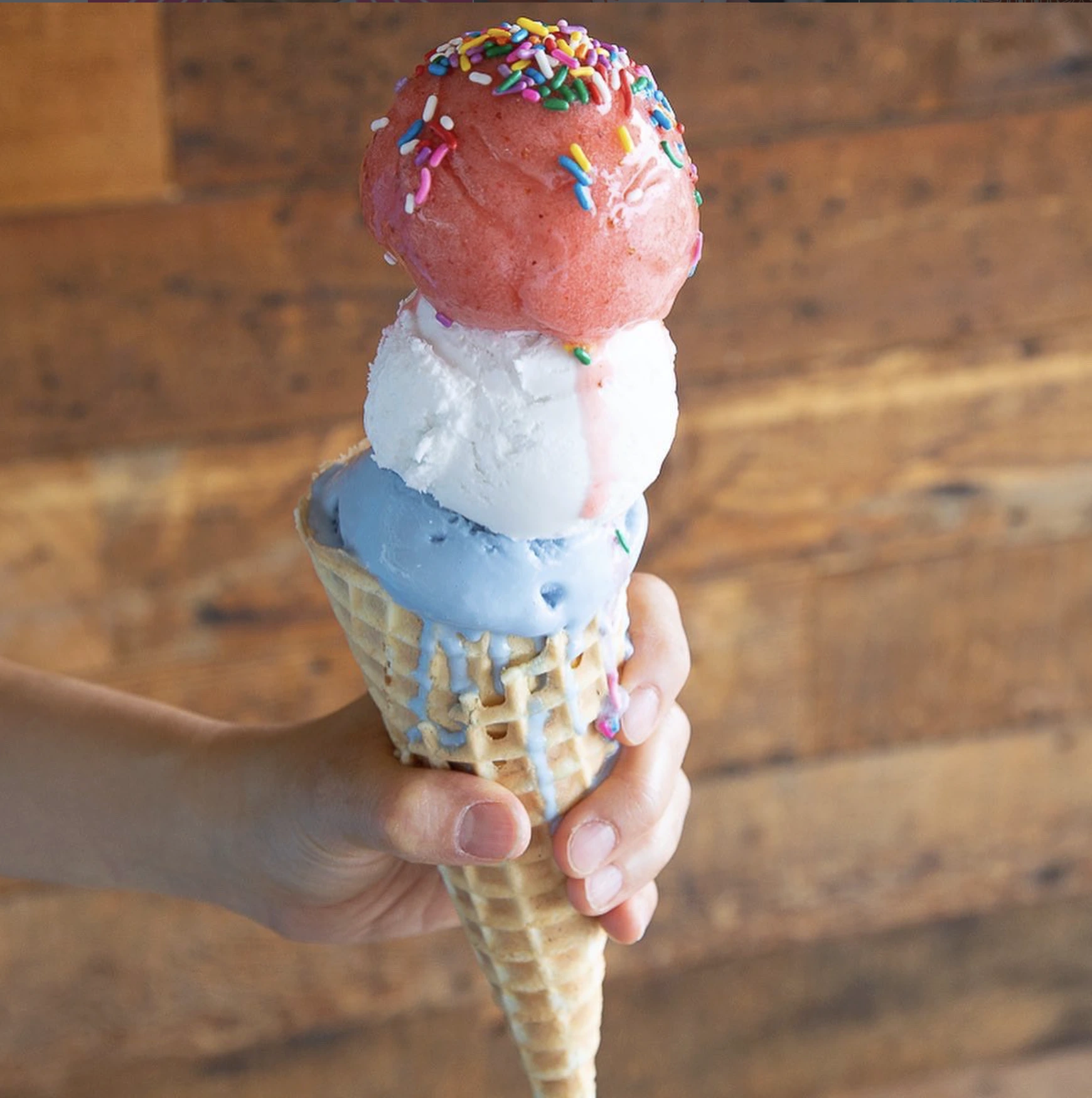 A hand holding a waffle cone with three scoops of Sweet Republic ice cream topped with sprinkles